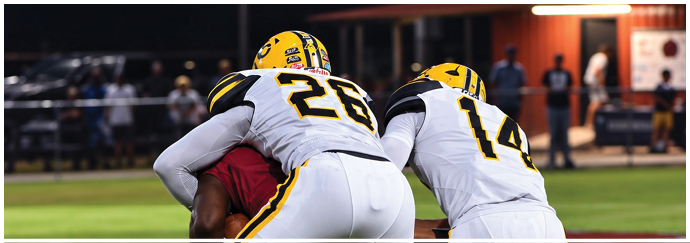 Two people in white and yellow sports uniforms are tackling a person in a red uniform on a grassy field during a game. Descripci n generada con IA