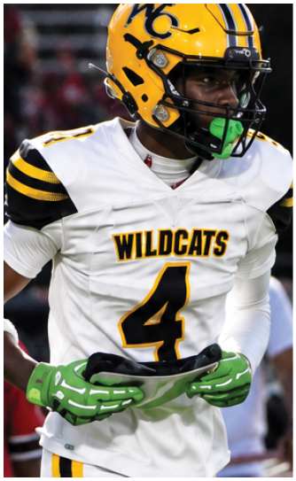 A person wearing a yellow and black helmet and white sports uniform is holding a green football on a field at night. Descripci n generada con IA