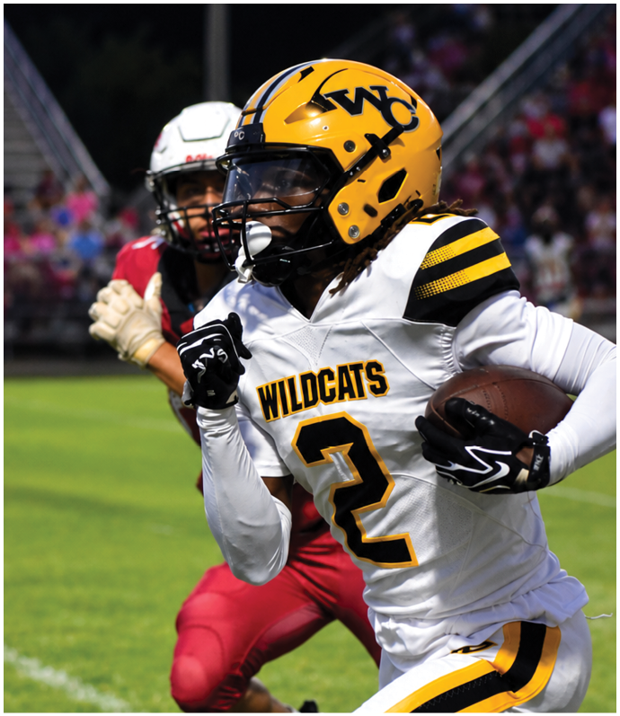 A person wearing a yellow helmet and white football uniform with “WILDCATS" and the number 2 is running with a football while being pursued by another person in a red uniform on a grassy field. Descripci n generada con IA