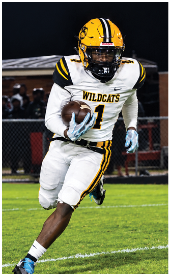 A person wearing a Wildcats football uniform and helmet is running on a grassy field at night while holding a football. Descripci n generada con IA