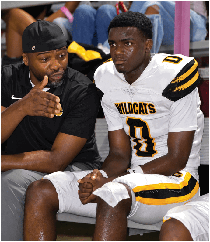 An adult wearing a black cap and shirt is talking and gesturing to a person in a white and yellow Wildcats football uniform sitting next to them on a bench. Descripci n generada con IA