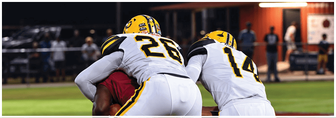 Two people in white and yellow sports uniforms are tackling a person in a red uniform on a grassy field during a game. Descripci n generada con IA