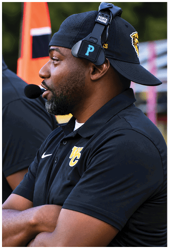 A person wearing a headset and a black polo shirt with a sports team logo stands with arms crossed, appearing focused on an event. Descripci n generada con IA
