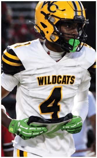 A person wearing a yellow and black helmet and white sports uniform is holding a green football on a field at night. Descripci n generada con IA