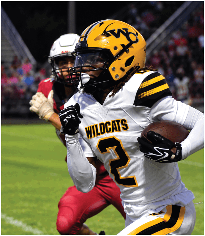 A person wearing a yellow helmet and white football uniform with “WILDCATS" and the number 2 is running with a football while being pursued by another person in a red uniform on a grassy field. Descripci n generada con IA