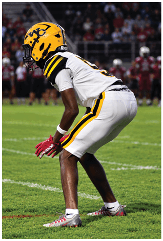 A person wearing a yellow football helmet and white uniform with black and yellow stripes is crouched on a grassy field, appearing ready to play. Descripci n generada con IA
