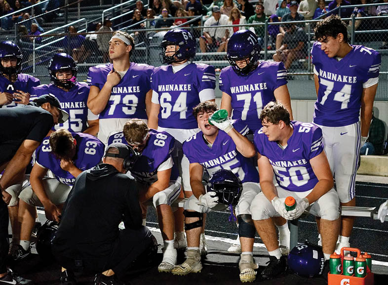 Four people wearing black football uniforms with “VISTA" on the front are standing together in a stadium with a crowd in the background, two of them holding helmets raised above their heads. Description generated by AI