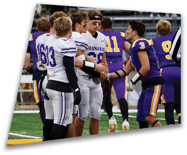 Four people in dark football uniforms and helmets stand in a row on a field, appearing focused and ready for action. Description generated by AI