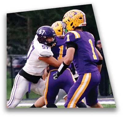 A person in football gear and helmet is crouched on a field with other players lined up in the background during a game. Description generated by AI