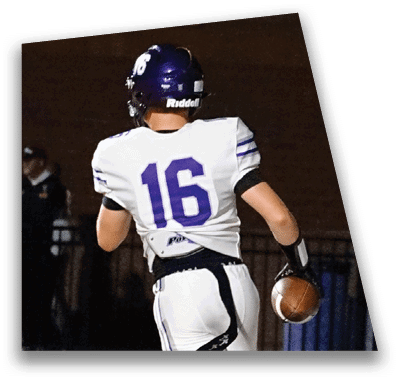 A person in football gear and helmet is crouched on a field with other players lined up in the background during a game. Description generated by AI