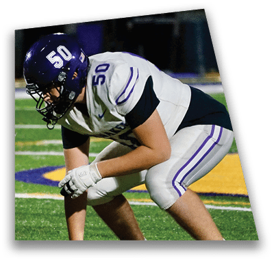 A person in football gear and helmet is crouched on a field with other players lined up in the background during a game. Description generated by AI