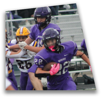 A person in football gear and helmet is crouched on a field with other players lined up in the background during a game. Description generated by AI