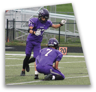 A person in football gear and helmet is crouched on a field with other players lined up in the background during a game. Description generated by AI