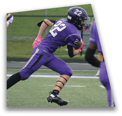 A person in football gear and helmet is crouched on a field with other players lined up in the background during a game. Description generated by AI