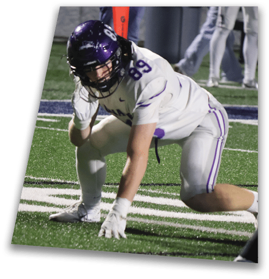 A person in a black football uniform and helmet is crouched in a ready position on a green field with another player in white uniform visible in the background. Description generated by AI