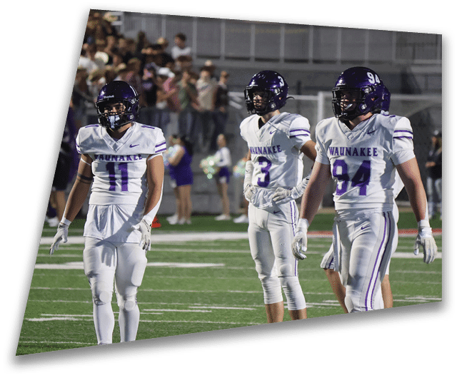 Four people in dark football uniforms and helmets stand in a row on a field, appearing focused and ready for action. Description generated by AI