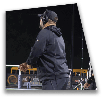 A person in football gear and helmet is crouched on a field with other players lined up in the background during a game. Description generated by AI