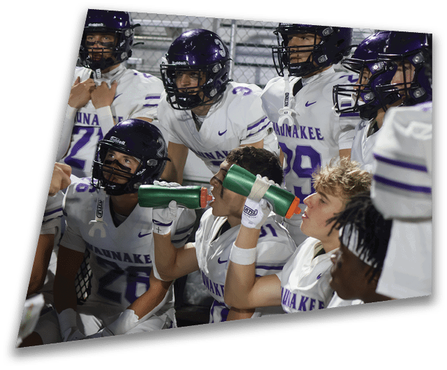 Four people in dark football uniforms and helmets stand in a row on a field, appearing focused and ready for action. Description generated by AI