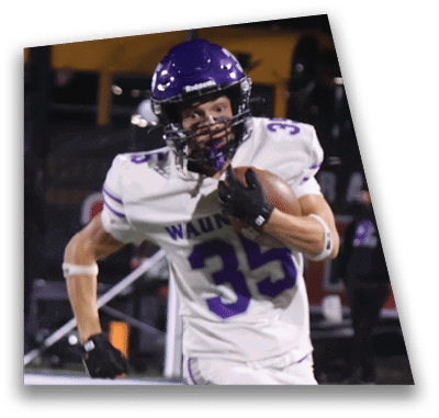 A person in football gear and helmet is crouched on a field with other players lined up in the background during a game. Description generated by AI