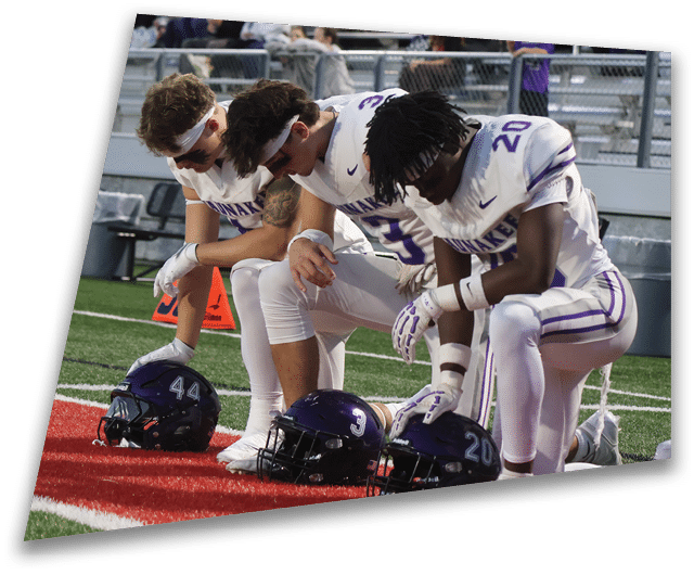 Four people in dark football uniforms and helmets stand in a row on a field, appearing focused and ready for action. Description generated by AI