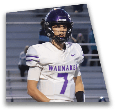A person in football gear and helmet is crouched on a field with other players lined up in the background during a game. Description generated by AI