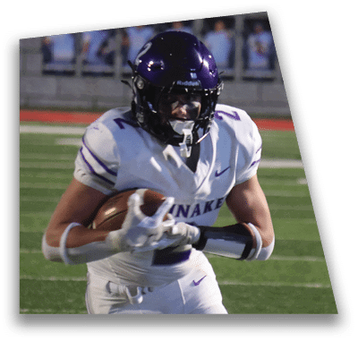A person in football gear and helmet is crouched on a field with other players lined up in the background during a game. Description generated by AI