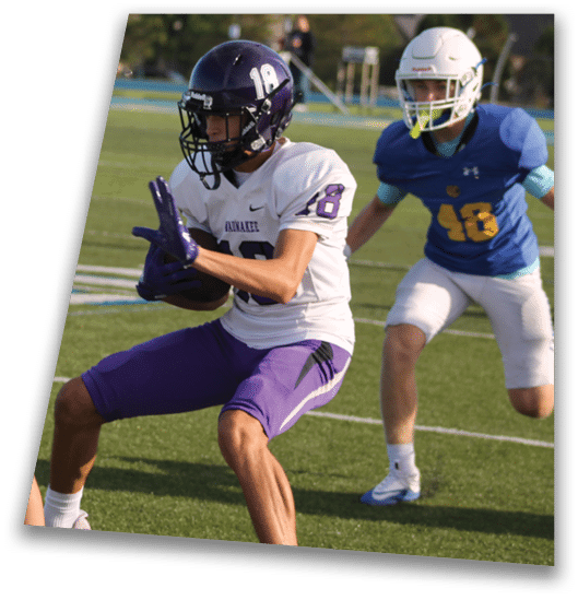 A person in a black football uniform and helmet is crouched in a ready position on a green field with another player in white uniform visible in the background. Description generated by AI