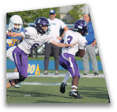 A person in football gear and helmet is crouched on a field with other players lined up in the background during a game. Description generated by AI