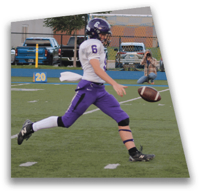 A person in football gear and helmet is crouched on a field with other players lined up in the background during a game. Description generated by AI