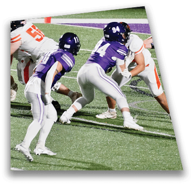 A person in football gear and helmet is crouched on a field with other players lined up in the background during a game. Description generated by AI