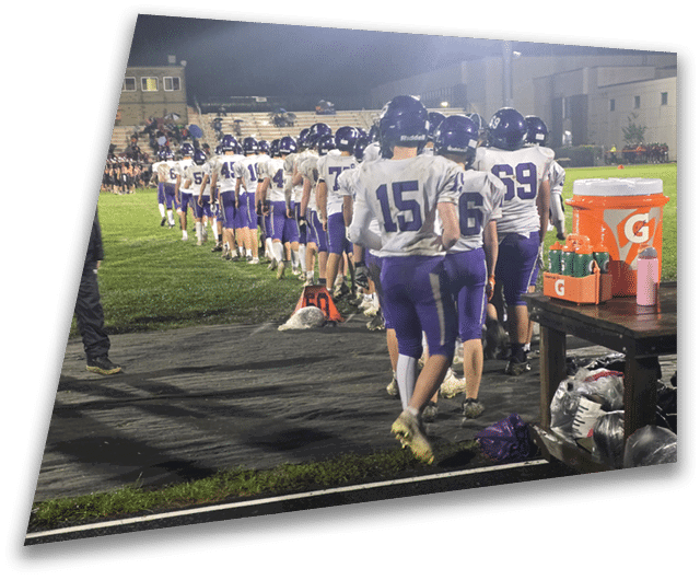 Four people in dark football uniforms and helmets stand in a row on a field, appearing focused and ready for action. Description generated by AI