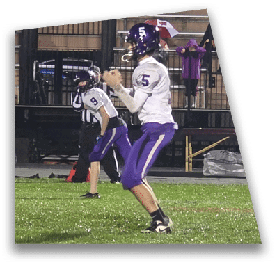 A person in football gear and helmet is crouched on a field with other players lined up in the background during a game. Description generated by AI