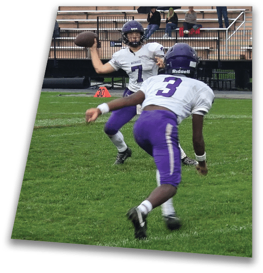 A person in a black football uniform and helmet is crouched in a ready position on a green field with another player in white uniform visible in the background. Description generated by AI