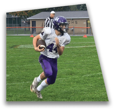 A person in football gear and helmet is crouched on a field with other players lined up in the background during a game. Description generated by AI