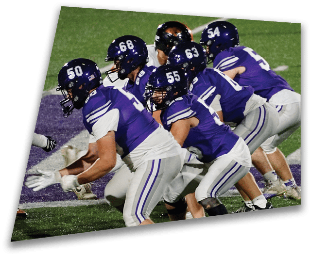 Four people in dark football uniforms and helmets stand in a row on a field, appearing focused and ready for action. Description generated by AI