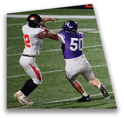 A person in football gear and helmet is crouched on a field with other players lined up in the background during a game. Description generated by AI