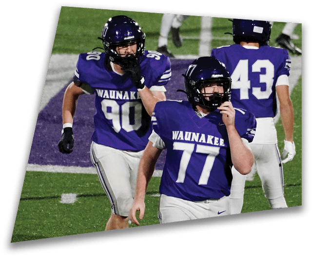 Four people in dark football uniforms and helmets stand in a row on a field, appearing focused and ready for action. Description generated by AI