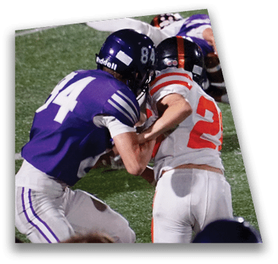 A person in football gear and helmet is crouched on a field with other players lined up in the background during a game. Description generated by AI
