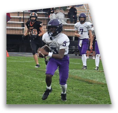 A person in football gear and helmet is crouched on a field with other players lined up in the background during a game. Description generated by AI