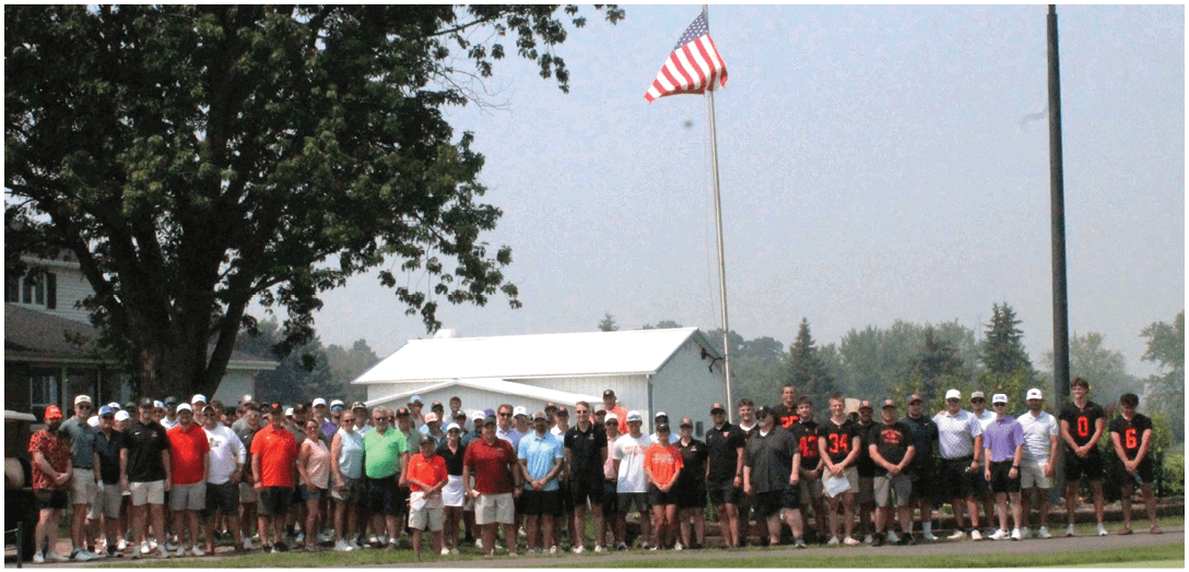 A large group of people wearing casual and sports clothing are standing outdoors in front of a white building and trees on a clear day. Description generated by AI