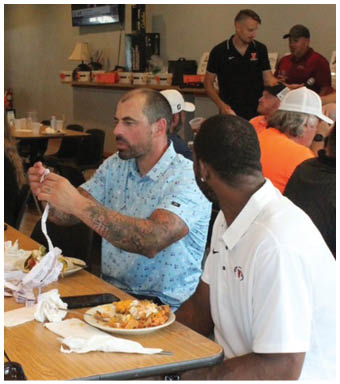 Two people sit at a table with plates of food while one person wearing a light blue patterned shirt with tattoos on their arms gestures with their hands. Description generated by AI