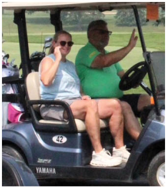 Two adults wearing shorts and sneakers are seated in a golf cart, with one person holding the steering wheel. Description generated by AI