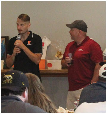Three people are gathered indoors with two wearing baseball caps and one holding a drink, with a bottle and other items visible in the background. Description generated by AI