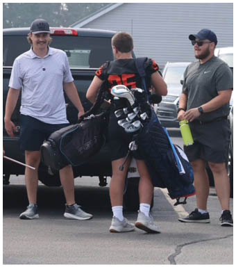 Two people wearing athletic shoes and shorts are standing near a vehicle, each carrying a golf bag. Description generated by AI