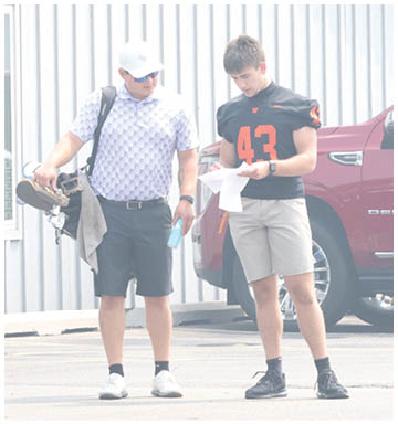 Two adults wearing shorts and watches stand near a vehicle while one holds a can and the other holds papers. Description generated by AI