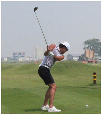 A person wearing a white cap and sleeveless shirt is swinging a golf club on a grassy field with buildings in the background. Description generated by AI