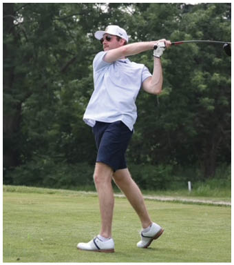 A person wearing a white cap, sunglasses, light blue shirt, navy shorts, and white golf shoes is swinging a golf club on a grassy area with trees in the background. Description generated by AI