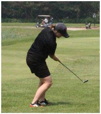 A person wearing a black outfit and cap is swinging a golf club on a grassy golf course with a golf cart and trees in the background. Description generated by AI