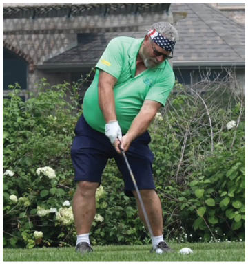 A person wearing a green shirt, navy shorts, and a star-spangled headband is preparing to hit a golf ball on a grassy area with bushes and a house in the background. Description generated by AI