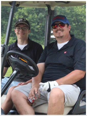 Two people wearing casual clothing and hats are sitting together in a golf cart with golf clubs visible in the background. Description generated by AI